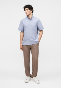 Άνδρας όρθιος φοράει γαλάζιο πουκάμισο με κοντά μανίκια και κουμπιά, καφέ παντελόνι και λευκά αθλητικά, κοιτάζοντας προς τα πλάγια.