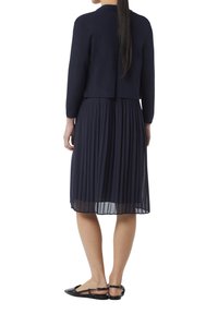 Marineblaues Plissékleid mit tailliertem Oberteil, langen Ärmeln und einer kurz geschnittenen Strickjacke. Flache schwarze Schuhe mit Knöchelriemen runden das Outfit ab.