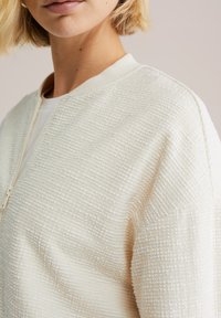 Krémovo sfarbená textúrovaná bunda so zipsovým zapínaním a rebrovaným golierom. Tkanina má vystúpený vzor a mierne voľný strih.