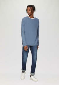Pull en maille texturée bleu avec un col rond, associé à un jean bleu foncé et des baskets blanches. Ce look allie confort et style décontracté.