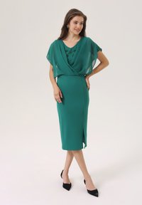 Robe turquoise avec une cape fluide et transparente superposée. Elle présente un décolleté en V et des embellissements décoratifs, longueur genou avec une fente sur le côté. Talons noirs.