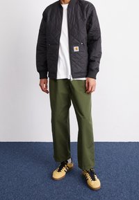 Sort quiltet bomberjakke med ribbede ærmeafslutninger, hvid t-shirt, olivengrønne viddebukser og gule sneakers med sorte accenter.