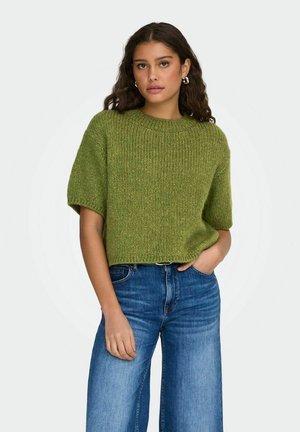 Giovane donna con lunghi capelli ricci che indossa un maglione verde a maniche corte lavorato a maglia e jeans blu a vita alta con gambe larghe, in piedi con una mano in tasca.