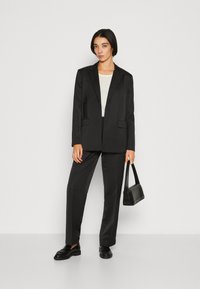 Black suit featuring a tailored blazer with notch lapels and matching trousers. Worn with a cream top and paired with a small black handbag.