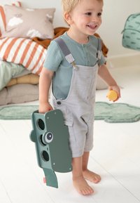 Groen speelgoed dat lijkt op een camera met gezichtskenmerken, vastgehouden door een kind dat lichtgrijze tuinbroek en een blauw geribd shirt draagt. Zachte binnenomgeving.