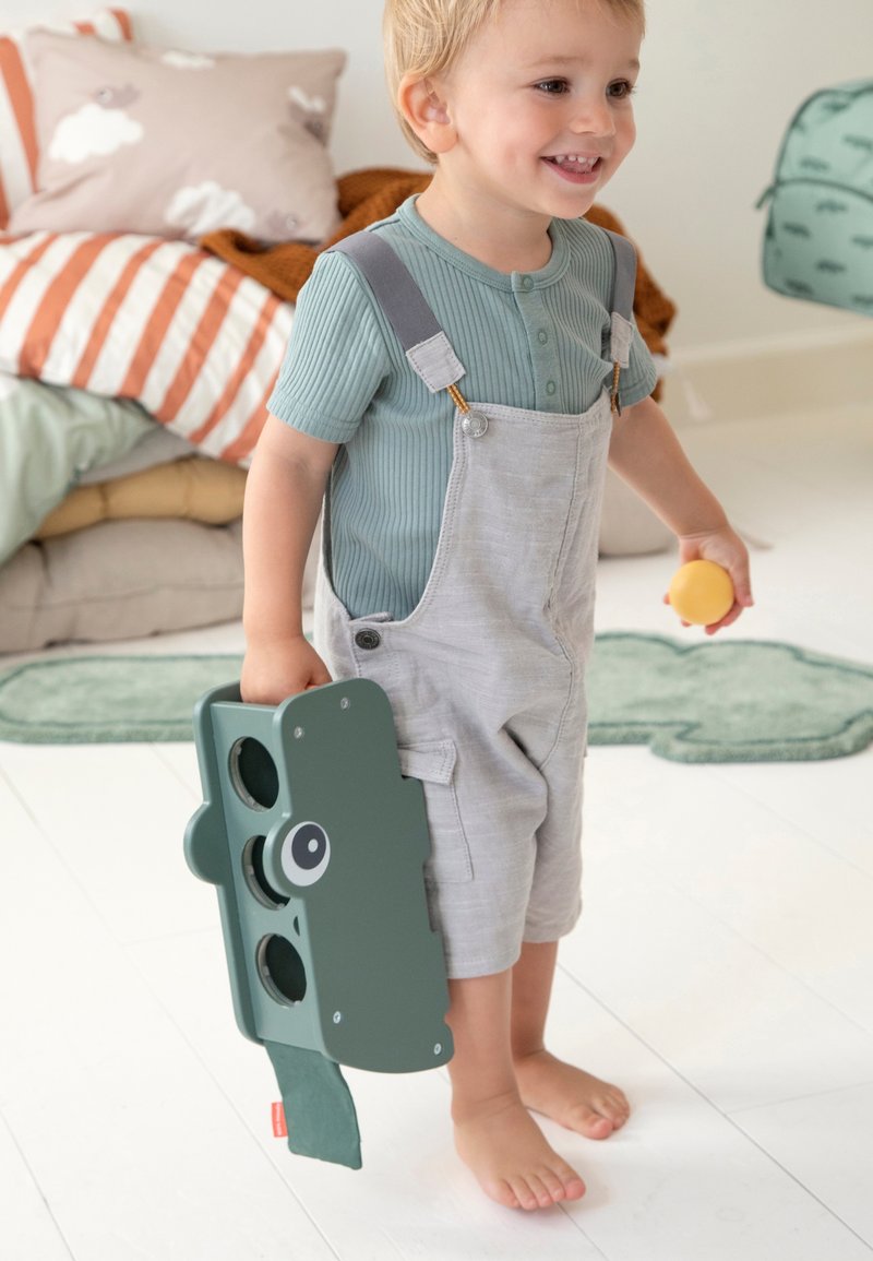 Groen speelgoed dat lijkt op een camera met gezichtskenmerken, vastgehouden door een kind dat lichtgrijze tuinbroek en een blauw geribd shirt draagt. Zachte binnenomgeving.