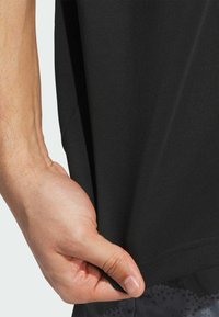 Sort atletisk t-shirt udstillet, som viser en glat stoftekstur, rund kant og subtile sømdetaljer. En hånd griber fat om kanten af t-shirten.