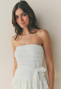 Robe blanche sans bretelles avec un tissu texturé présentant des ondulations horizontales et un ourlet froncé, cintrée à la taille par une ceinture nouée.