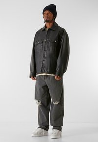 Schwarze Lederjacke mit zwei vorderen Taschen, getragen über einem cremefarbenen Hemd. Zerrissene graue Jeans und weiße Sneakers runden das Outfit ab.