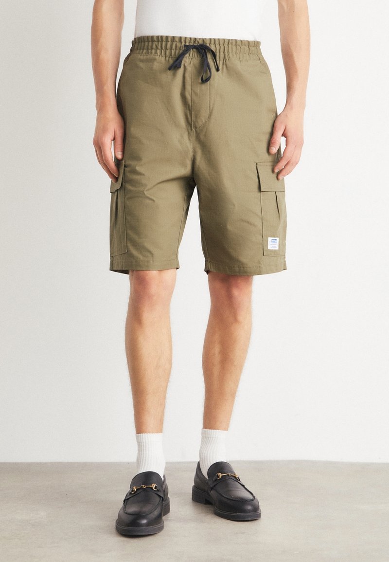 Homme portant un short cargo vert olive avec taille à cordon, chaussettes blanches et mocassins en cuir noir debout sur un sol en béton.