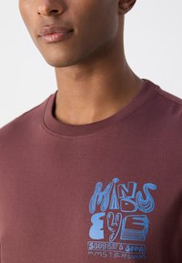 Bordová bavlnen�á tričko s modrým grafickým textom, ktorý znie "MIND'S EYE SCOTCH & SODA AMSTERDAM" na hrudi.