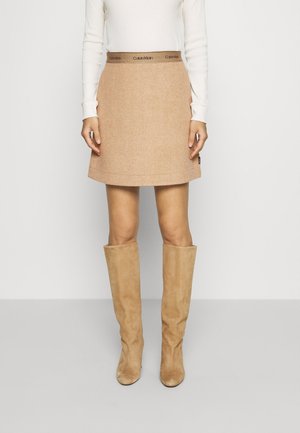 Vrouw die een beige minirok draagt met een Calvin Klein tailleband, een wit top met lange mouwen en beige, kniehoge suède laarzen, staande tegen een witte muur.