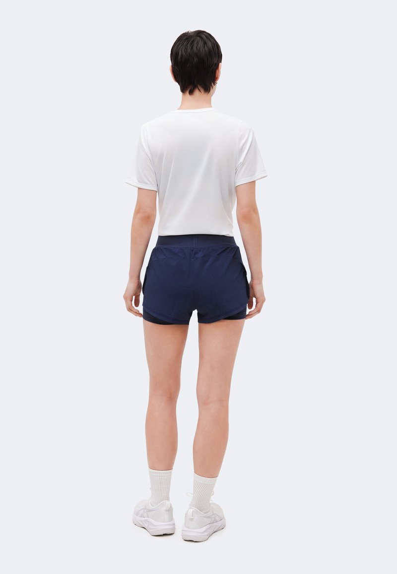 Person steht mit dem Rücken zur Kamera, trägt ein weißes kurzärmeliges Hemd, marineblaue Sportshorts, weiße Socken und weiße Turnschuhe vor einem einfarbigen Hintergrund.