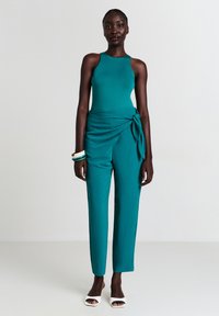 Combinaison en bleu sarcelle avec un haut sans manches ajusté et un pantalon large, présentant un nœud décoratif sur le côté. Tissu lisse et design minimaliste.