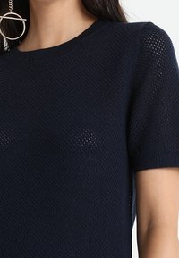 Haut tricoté à manches courtes bleu marine avec un col rond, un motif légèrement ouvert et des bords côtelés pour ajouter de la texture et favoriser la respirabilité.