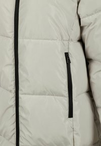 Vaaleanbeige puffatakki, jossa on musta etuvetoketju ja vetoketjullinen sivutasku, sekä tikkaukset yksityiskohtina.