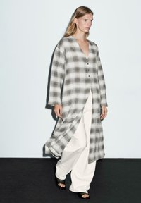 Lekka, długa koszula z guzikami w szaro-białą kratę. Posiada długie rękawy, głęboki dekolt w serek oraz boczne rozcięcia. Zestawiona z białymi spodniami o szerokich nogawkach.