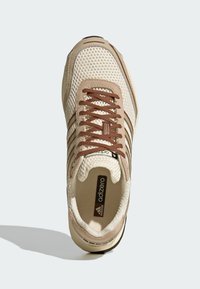 Μπεζ αθλητικό παπούτσι με άνω μέρος από δίχτυ, καφέ κορδόνια και τρεις σκουρότερες καφέ ρίγες. Η σόλα είναι στρογγυλεμένη και έχει υφή με ανάγλυφο σχέδιο.