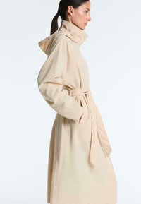Femme en trench-coat beige avec col montant et capuche, les mains dans les poches, vue de profil sur un fond uni.