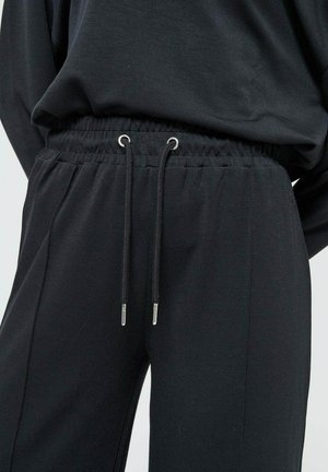 Un pantalon de jogging noir doté d'une taille élastique avec cordons de serrage ajustables et embouts en métal, fabriqué à partir d'un tissu doux et texturé.