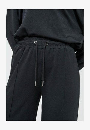 Un pantalon de jogging noir doté d'une taille élastique avec cordons de serrage ajustables et embouts en métal, fabriqué à partir d'un tissu doux et texturé.