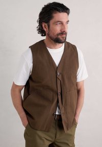 Brązowa bawełniana kamizelka z dwoma przednimi kieszeniami, zapinana na guziki, noszona na białej koszulce. Model stoi z rękami w kieszeniach.