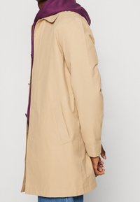 Persona che indossa un trench beige sopra un felpa viola con le mani incrociate dietro la schiena, su uno sfondo bianco uniforme.