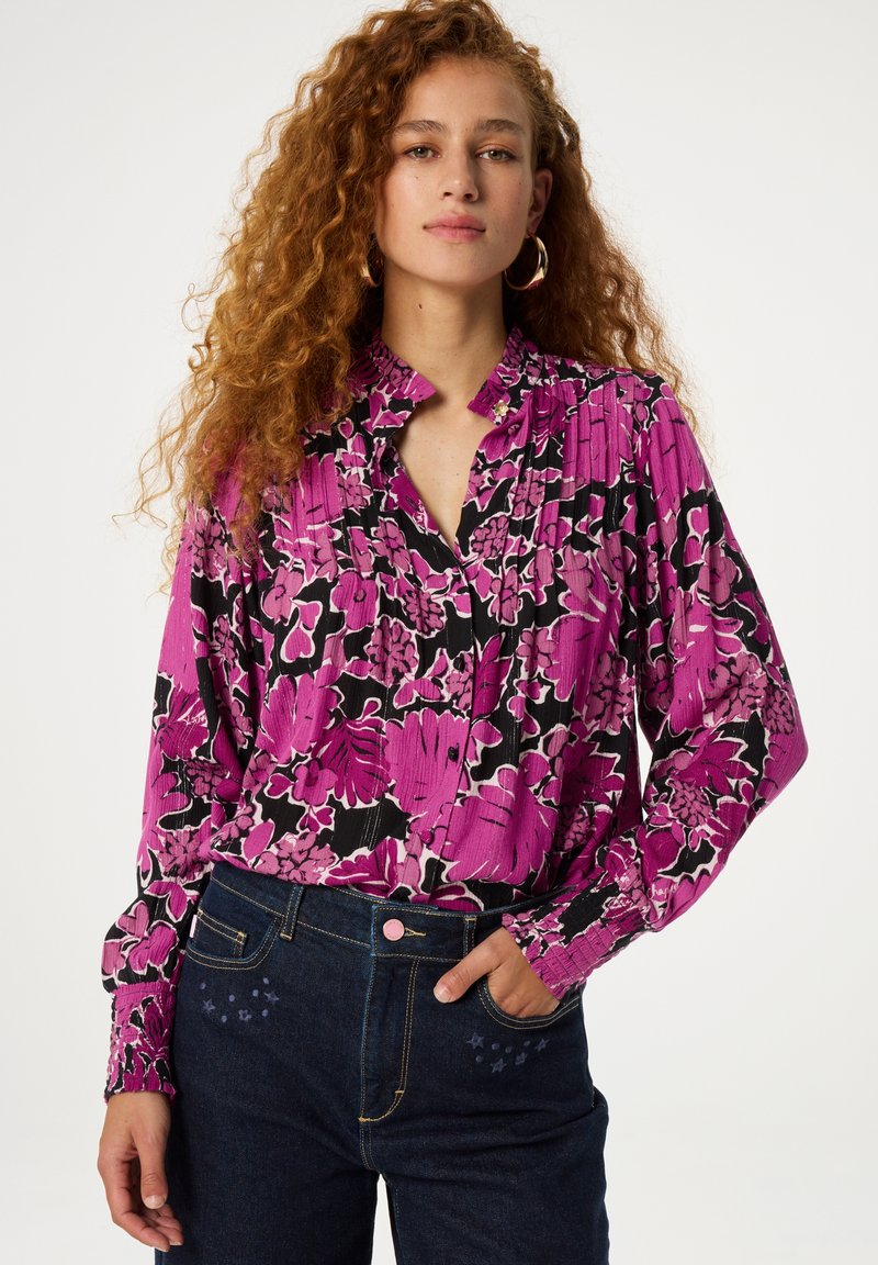 Pink floral blouse with a pleated texture, featuring a ruffled collar and black accents, paired with dark denim jeans and a pink button.