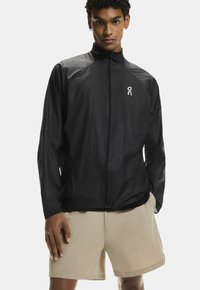 Melns viegls jaka ar augstu apkakli, priekšējo rāvējslēdzēju un atstarojošu logo. Iespējas ar garām piedurknēm un gludu, plānu auduma tekstūru.