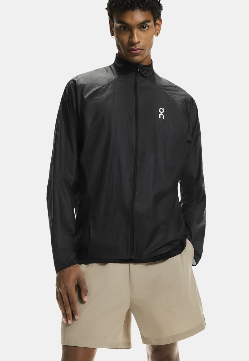 Melns viegls jaka ar augstu apkakli, priekšējo rāvējslēdzēju un atstarojošu logo. Iespējas ar garām piedurknēm un gludu, plānu auduma tekstūru.