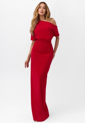 Kobieta w długiej czerwonej sukni z odsłoniętymi ramionami stojąca na jednolitym białym tle, z luźnymi falami włosów i minimalistyczną biżuterią.