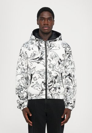 Young man wearing a white zip-up hooded jacket with black and gray ornate floral patterns and black pants, standing against a plain background.