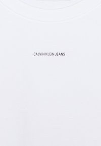 White cotton t-shirt featuring a crew neck, ribbed texture, and a small gray "CALVIN KLEIN JEANS" logo centered on the chest.