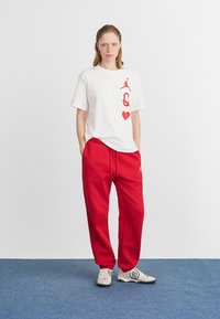 Camiseta blanca con logo gráfico rojo y corazón. Pantalones de chándal rojos con cinturilla elástica. Zapatillas grises con detalles negros y logo. Suelo azul.