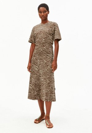 Frau mit kurzen Haaren, die ein braunes und beiges Midikleid mit Zebramuster sowie braune Sandalen mit Knöchelriemen trägt, steht vor einem weißen Hintergrund.