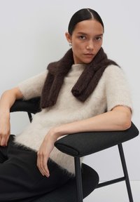 Un pull en tricot beige clair à manches courtes, avec un plaid en peluche marron foncé. Assise sur une chaise noire, avec des cheveux lisses et un maquillage naturel.