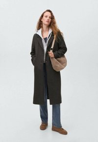 Woman with long curly hair wearing a dark long coat over a gray hoodie, blue jeans, brown shoes, and carrying a beige shoulder bag.