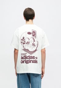 Kremkrāsas kokvilnas T-krekls ar bordo grafiku uz aizmugures, attēlojot krēslu un augus, ar tekstu "adidas originals".