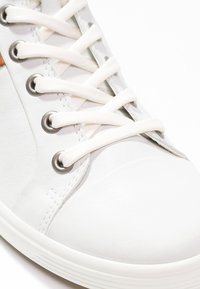 Weißer Sneaker mit einem strukturierten Obermaterial aus synthetischem Leder, grauen Ösen, flachen weißen Schnürsenkeln und einer Gummisohle. Präsentiert ein minimalistisches Design.