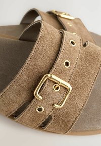 Sandales en suède marron avec deux larges brides, dotées de boucles dorées et d'œillets argentés. Matériau texturé et design à bout ouvert.