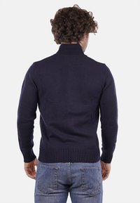 Maglione in navy a maglia con colletto alto, polsini e orlo a costine. Presenta un design aderente e un tessuto testurizzato. Jeans in denim blu visibili.