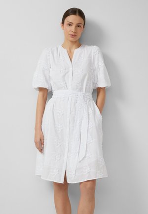 Kobieta ubrana w białą sukienkę do kolan z bufkami, kwiatowym haftem, zapinana na guziki z przodu i przewiązanym paskiem w talii, stoi przy szarej ścianie.