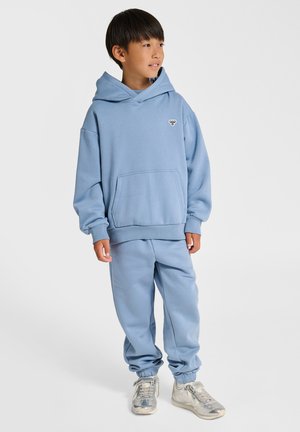 Lyseblå hættetrøje med frontlomme og logo, kombineret med matchende joggingbukser. Stoffet er blødt med en glat tekstur. Sølvfarvede sneakers vises.