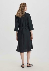 Abito in lino nero con maniche a tre quarti, vita cintata e spacco laterale. Presenta una vestibilità morbida e una texture delicata. Sandali completano l'outfit.