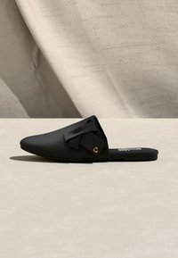 Μαύρο μοκασίνι με επίπεδο πάτο και λάστιχο, μυτερή μύτη, διακοσμητικό φιόγκο και μικρό χρυσό καρδιά στην ουδέτ ερη φόντο.