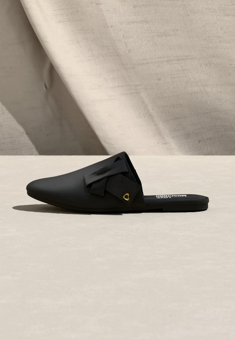 Mocassin plat noir à enfiler avec un bout pointu, un nœud décoratif et un petit charm en forme de cœur doré sur un fond neutre.