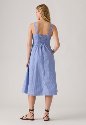 Woman with long blonde hair wearing a blue and white vertical striped sleeveless midi dress and black lace-up sandals, standing facing away.