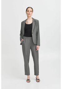 Costume gris ajusté comprenant un blaser cintré et un pantalon fuselé. Le tissu semble lisse. Associé à un top noir et des sandales à lanières.