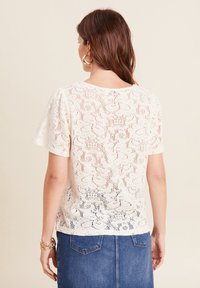 Top in pizzo color crema con maniche corte e motivi floreali, dotato di un collo rotondo e una vestibilità leggermente ampia. Indossato con una gonna di jeans.
