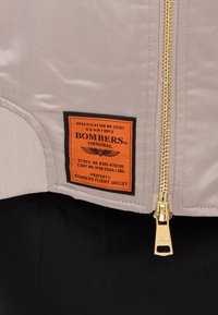 Sezione di giacca bomber color ruggine con cerniera dorata ed etichetta rettangolare arancione con testo nero ed emblema alato.